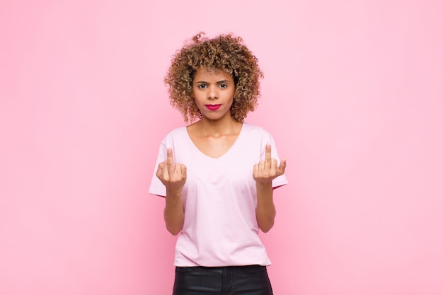 Joven mujer afroamericana sintiéndose provocativa, agresiva y obscena, moviendo el dedo medio, con una actitud rebelde contra la pared rosa