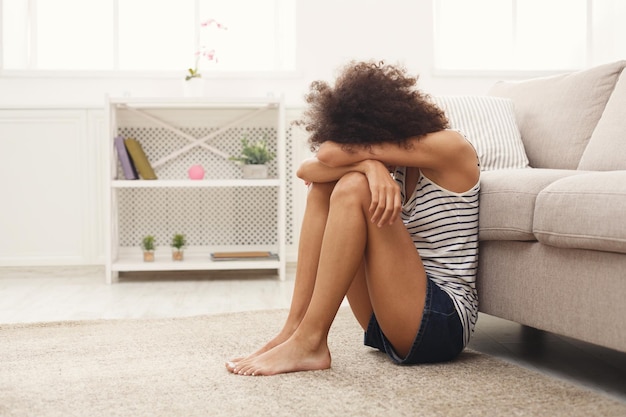 Joven mujer afroamericana solitaria sentada en el suelo en casa. Chica pensativa y triste que se esconde del mundo, copie el espacio. Depresión, concepto de estrés