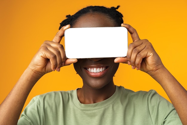 Joven mujer afroamericana sonriente que muestra un teléfono inteligente con pantalla en blanco contra un fondo amarillo