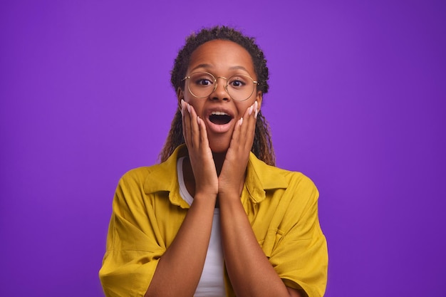 Joven mujer afroamericana sorprendida gen z abriendo la boca pone las manos en las mejillas