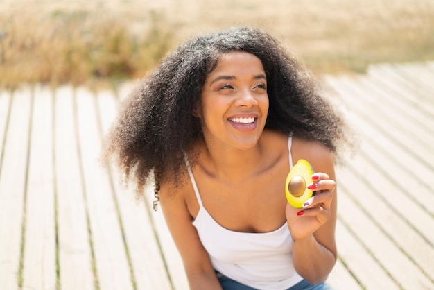 Joven mujer afroamericana sosteniendo un aguacate al aire libre mirando hacia arriba mientras sonríe