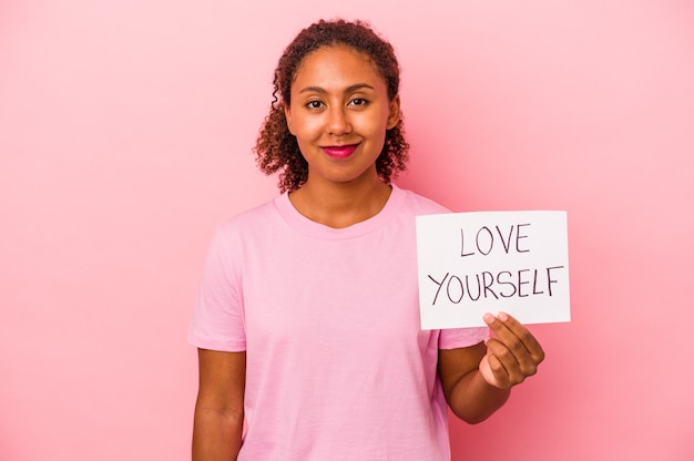 Joven mujer afroamericana sosteniendo ámate a ti mismo cartel aislado sobre fondo de color rosa