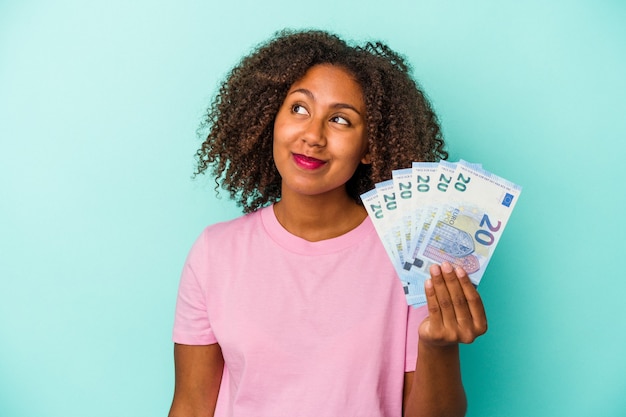 Joven mujer afroamericana sosteniendo billetes en euros aislado sobre fondo azul soñando con lograr metas y propósitos