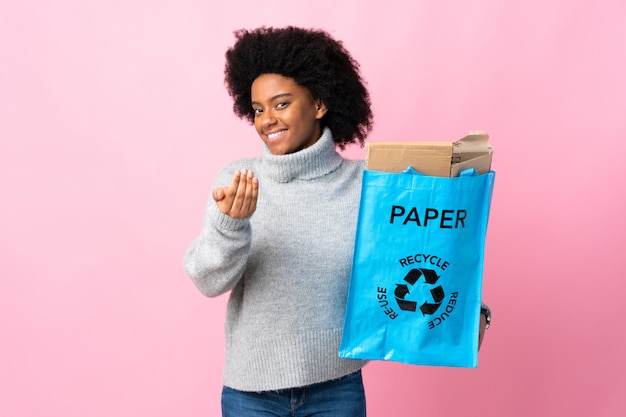 Joven mujer afroamericana sosteniendo una bolsa de reciclaje aislada en coloridos invitando a venir con la mano. Feliz de que hayas venido