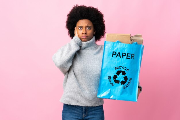 Joven mujer afroamericana sosteniendo una bolsa de reciclaje aislada sobre fondo colorido frustrado y cubriendo las orejas