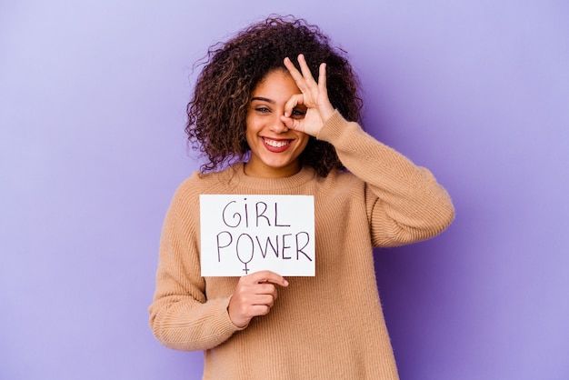 Foto joven mujer afroamericana sosteniendo un cartel de girl power en púrpura emocionado manteniendo el gesto ok en el ojo.