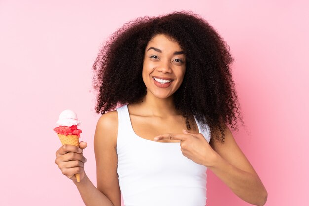 Joven mujer afroamericana sosteniendo un helado de cucurucho aislado en rosa y señalando