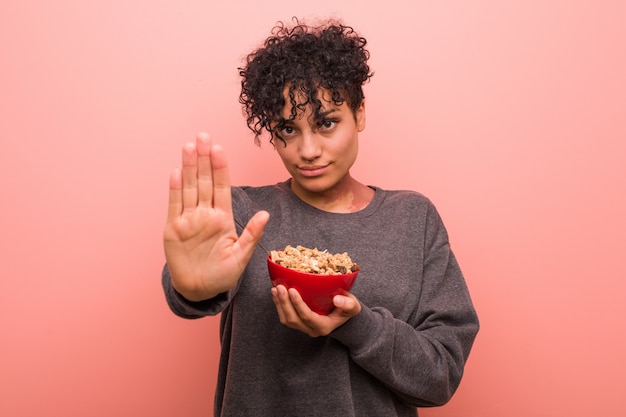 Joven mujer afroamericana sosteniendo un tazón de cereal de pie con la mano extendida que muestra la señal de stop, impidiéndole.