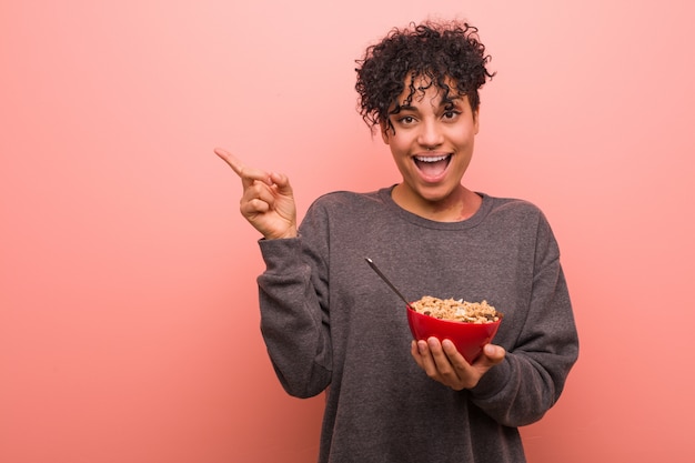 Joven mujer afroamericana sosteniendo un tazón de cereal sonriendo alegremente señalando con el dedo lejos.