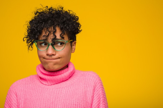 Joven mujer afroamericana con un suéter rosa confundido, se siente dudoso e inseguro.