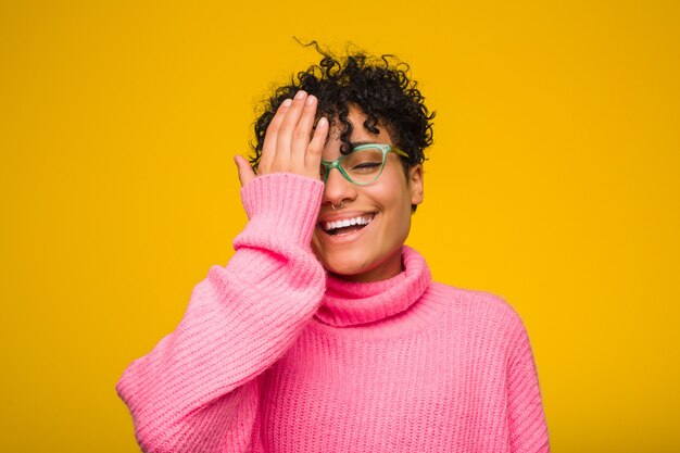 Joven mujer afroamericana con un suéter rosa divirtiéndose cubriendo la mitad de la cara con la palma.