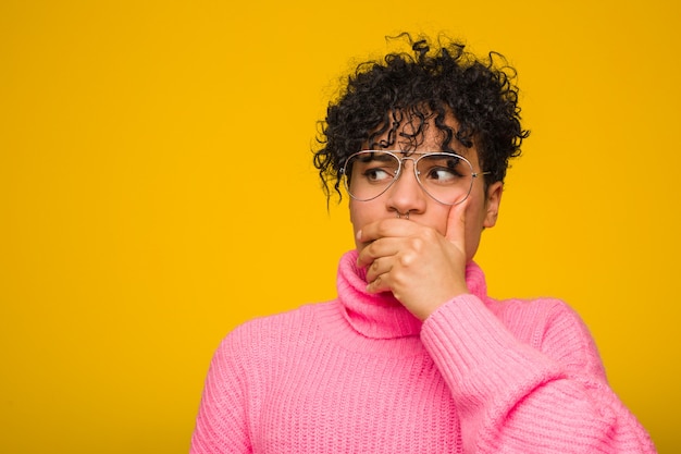 Joven mujer afroamericana con un suéter rosa pensativo a una boca que cubre con la mano.