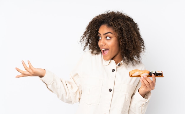 Joven mujer afroamericana con sushi en la pared blanca con expresión facial sorprendida