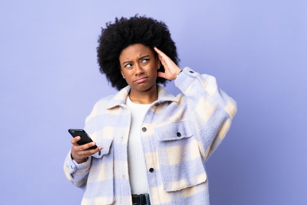 Joven mujer afroamericana utilizando teléfono móvil aislado sobre fondo púrpura con dudas y con expresión de la cara confusa