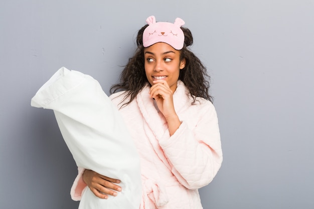 Joven mujer afroamericana vistiendo un pijama y una máscara para dormir con una almohada relajada pensando en algo mirando un espacio de copia.