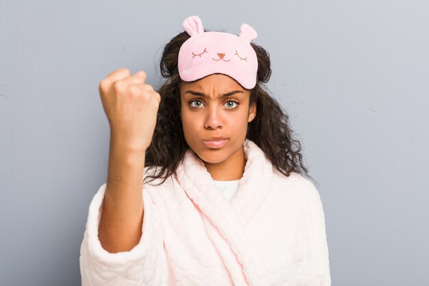 Joven mujer afroamericana vistiendo un pijama y una máscara para dormir mostrando el puño a la cámara, expresión facial agresiva.