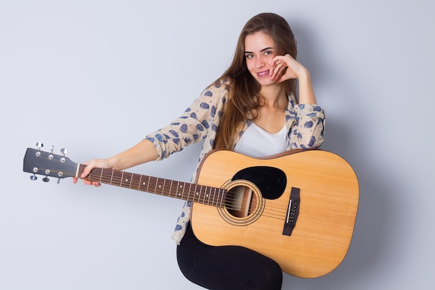 Joven mujer agradable con blusa beige y pantalones negros sosteniendo una guitarra con fondo gris en el estudio