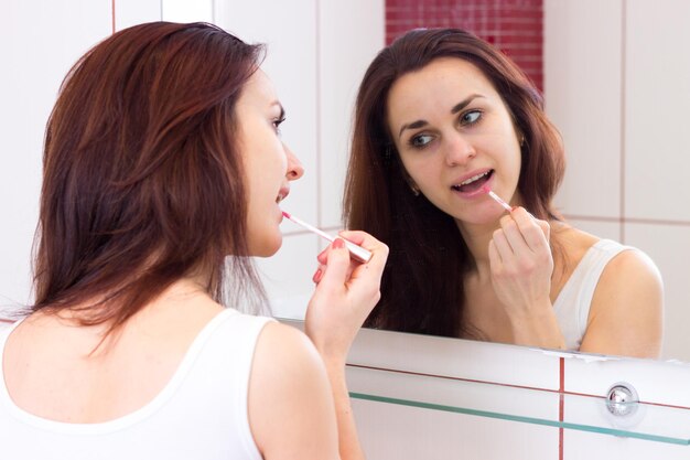 Joven mujer agradable con brillo de labios de camisa frente al espejo en su baño de color burdeos