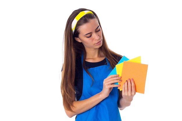 Joven mujer agradable con camiseta azul y delantal sosteniendo plumeros sobre fondo blanco en el estudio