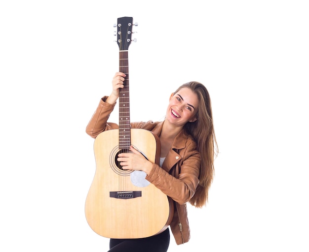 Joven mujer agradable con chaqueta marrón sosteniendo una guitarra con fondo blanco en el estudio