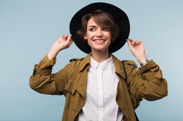 Joven mujer alegre con cabello corto oscuro en gabardina y sombrero felizmente mirando a un lado sobre fondo azul aislado