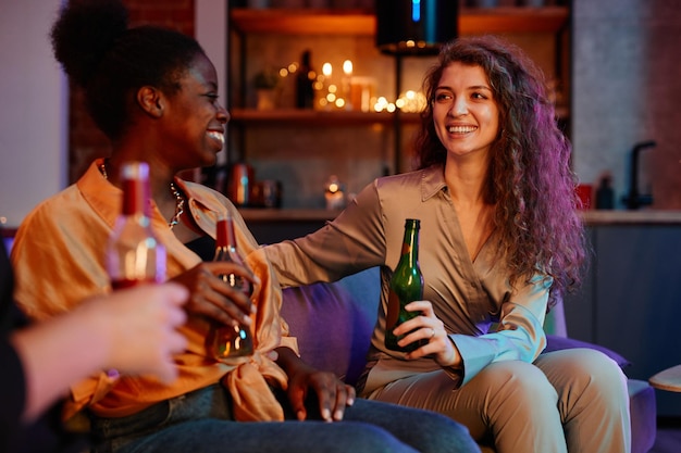 Joven mujer alegre con cabello largo y grueso tomando cerveza y hablando con una novia afroamericana sentada