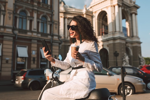 Joven mujer alegre con cabello rizado oscuro con gafas de sol y traje blanco sentada en un ciclomotor con una taza de café para llevar y felizmente usando su teléfono celular con vista a la ciudad en el fondo