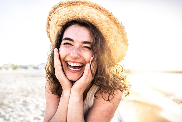 Joven mujer alegre con camisa blanca y sombrero sonriendo a la cámara en la playa