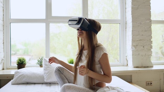 Joven mujer alegre con casco de realidad virtual sentada en la cama en casa