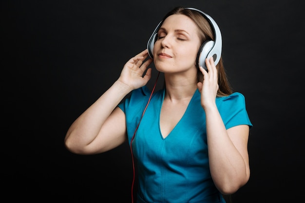 Joven mujer alegre positiva que se siente en paz y usa auriculares mientras disfruta de la música y de pie contra la pared negra