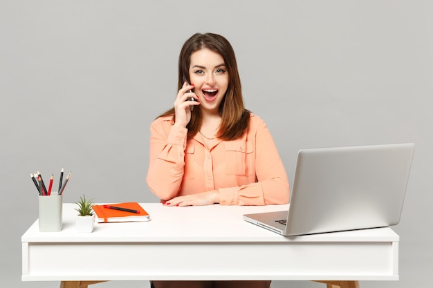 Joven mujer alegre con ropa informal en colores pastel hablando por teléfono móvil, siéntese a trabajar en el escritorio con una computadora portátil aislada en un fondo gris. Concepto de estilo de vida de carrera empresarial de logro. Simulacros de espacio de copia.