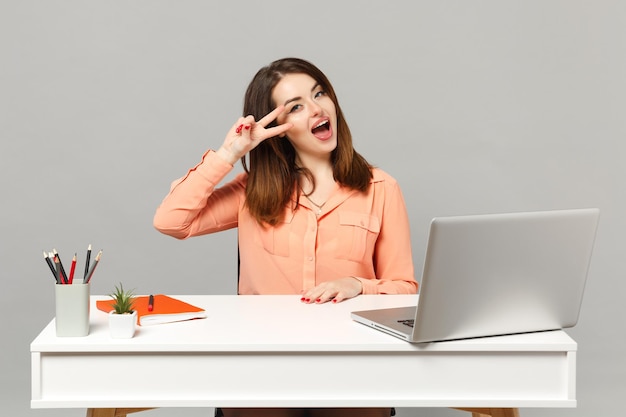 Una joven mujer alegre con ropa informal en colores pastel que muestra el signo de la victoria se sienta a trabajar en un escritorio blanco con una computadora portátil aislada en un fondo gris. Concepto de estilo de vida de carrera empresarial de logro. Simulacros de espacio de copia.