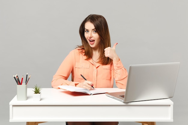 Joven mujer alegre con ropa pastel que muestra el pulgar hacia arriba, escribe notas, se sienta a trabajar en el escritorio con una computadora portátil aislada en un fondo gris. Concepto de estilo de vida de carrera empresarial de logro. Simulacros de espacio de copia.