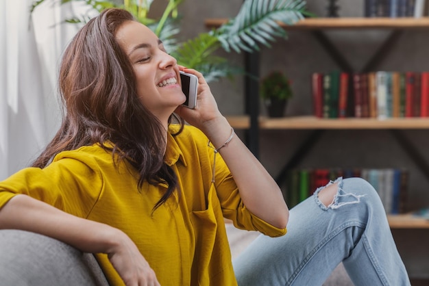 Joven mujer alegre sentada en el sofá y hablando por teléfono móvil en casa