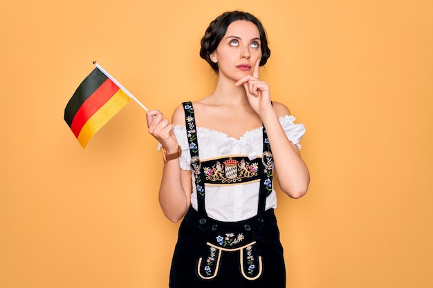 Joven mujer alemana patriótica con ojos azules que lleva un vestido de Oktoberfest sosteniendo la bandera de Alemania cara seria pensando en la pregunta idea muy confusa