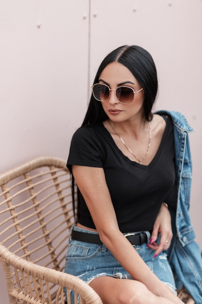 Joven mujer americana con pelo largo con gafas de sol en una elegante chaqueta de mezclilla con una camiseta negra sentada en una silla cerca de una pared rosa en la terraza de un café de verano. Chica con estilo sexy hipster al aire libre.