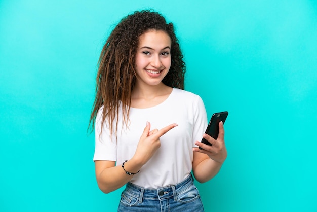 Joven mujer árabe aislada de fondo azul usando un teléfono móvil y apuntándolo