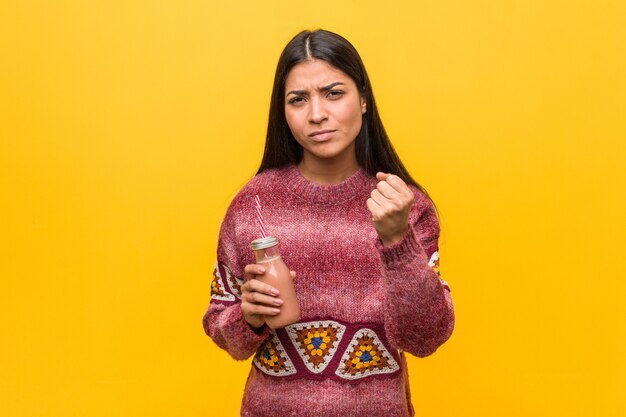 Joven mujer árabe sosteniendo un batido mostrando el puño a la cámara, expresión facial agresiva.