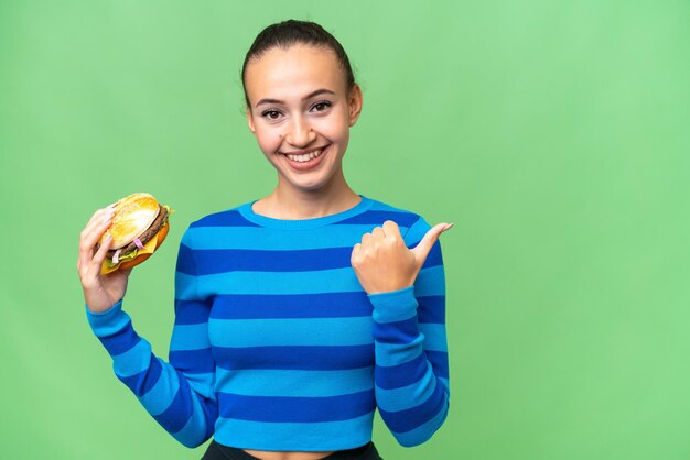 Joven mujer árabe sosteniendo una hamburguesa sobre un fondo aislado apuntando hacia un lado para presentar un producto