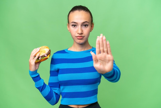 Joven mujer árabe sosteniendo una hamburguesa sobre un fondo aislado haciendo un gesto de parada