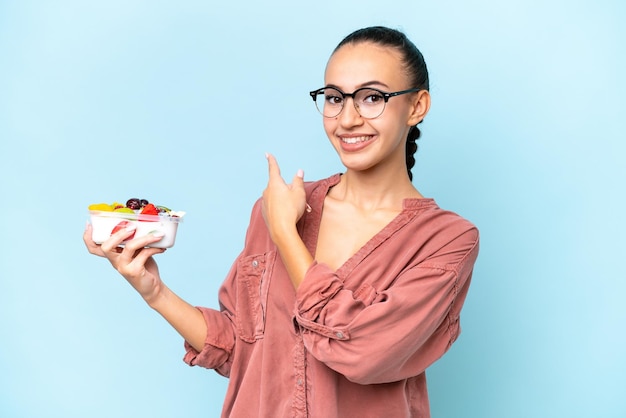 Joven mujer árabe sosteniendo un plato de fruta aislado sobre fondo azul apuntando hacia atrás