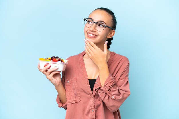 Joven mujer árabe sosteniendo un tazón de fruta aislada de fondo azul mirando hacia arriba mientras sonríe