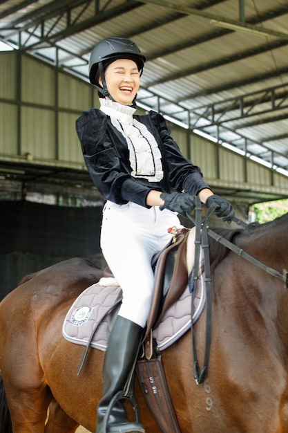 Joven mujer asiática de 20 años con traje de montar a caballo con un hermoso animal al aire libre. Paseo femenino alto, inteligente, fuerte, caballo, en, estable, y, entrene, aprendizaje, práctica, encima, fondo verde, longitud completa