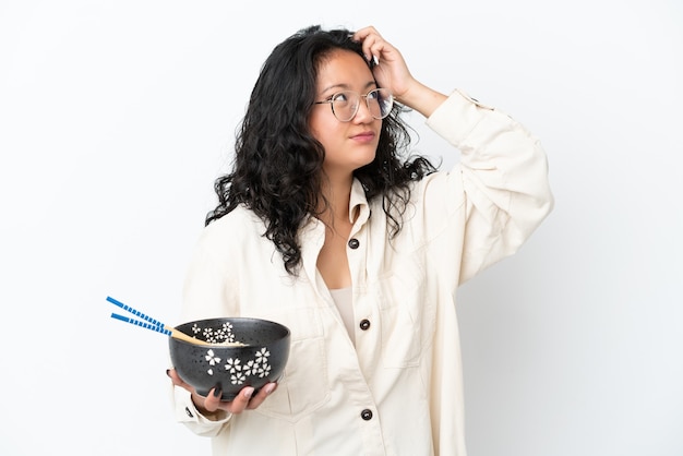 Joven mujer asiática aislada sobre fondo blanco que tiene dudas y confunde la expresión de la cara mientras sostiene un plato de fideos con palillos
