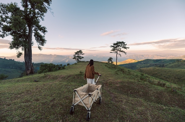 Joven mujer asiática arrastrando carro de camping en la colina verde en el campo al atardecer