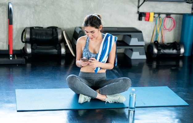 Joven mujer asiática atractiva sonriente fitness activo sentado en el suelo del gimnasio y con smartphone