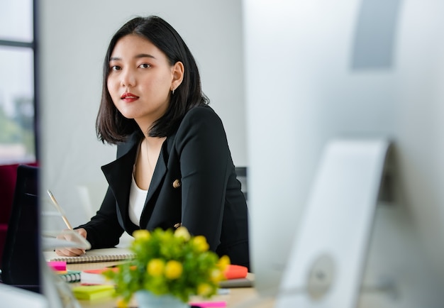 Joven mujer asiática atractiva en traje negro sentado en la oficina moderna trabajando y sonriendo con el portátil sobre la mesa. Concepto de estilo de vida de oficina moderna.