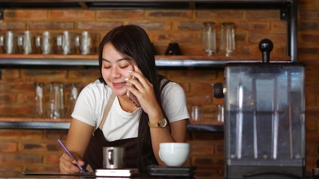 Joven mujer asiática barista usa delantal hablando y recibe el pedido del cliente en el teléfono celular en la cafetería. Concepto de pequeña empresa de tienda de café. Camarero femenina escribiendo una nota mientras escucha al cliente