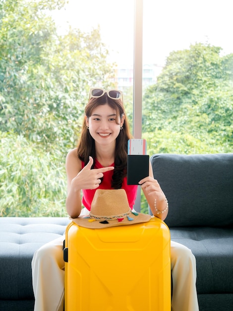 Joven mujer asiática bastante feliz con gafas de sol que señalan el pasaporte y el boleto de avión con una sonrisa sentada en el sofá cerca del sombrero de playa maleta amarilla estilo vertical lista para viajar en concepto de vacaciones de verano