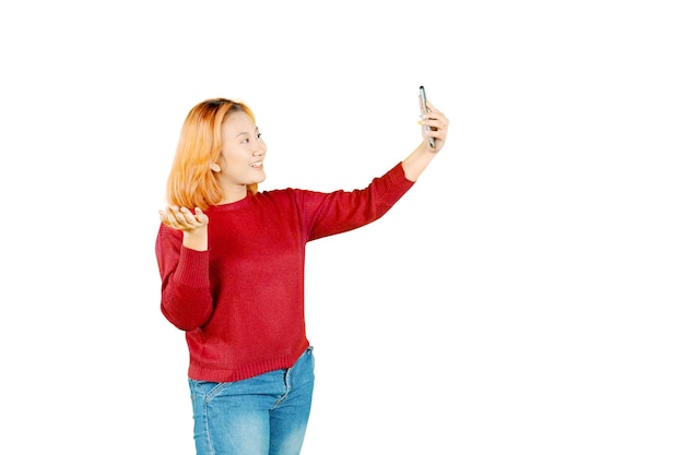 Joven mujer asiática bonita de pie haciendo un selfie disparado en el teléfono móvil saludando con la mano aislada sobre fondo blanco